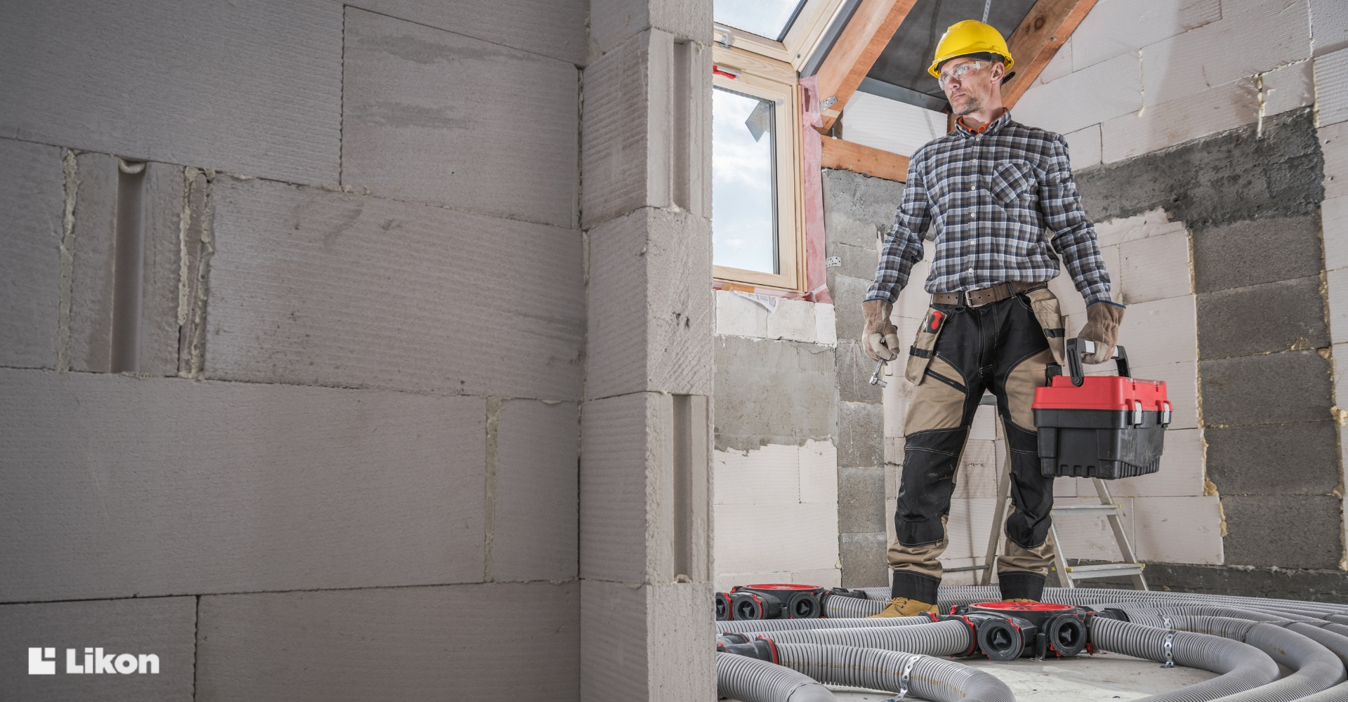Instalator w kasku ochronnym stoi w budowanym domu obok rozprowadzonych rur systemu rekuperacji, trzymając skrzynkę z narzędziami.