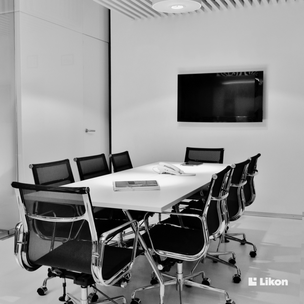 Nowoczesna sala konferencyjna z długim stołem, krzesłami na kółkach oraz ekranem multimedialnym zamontowanym na ścianie.