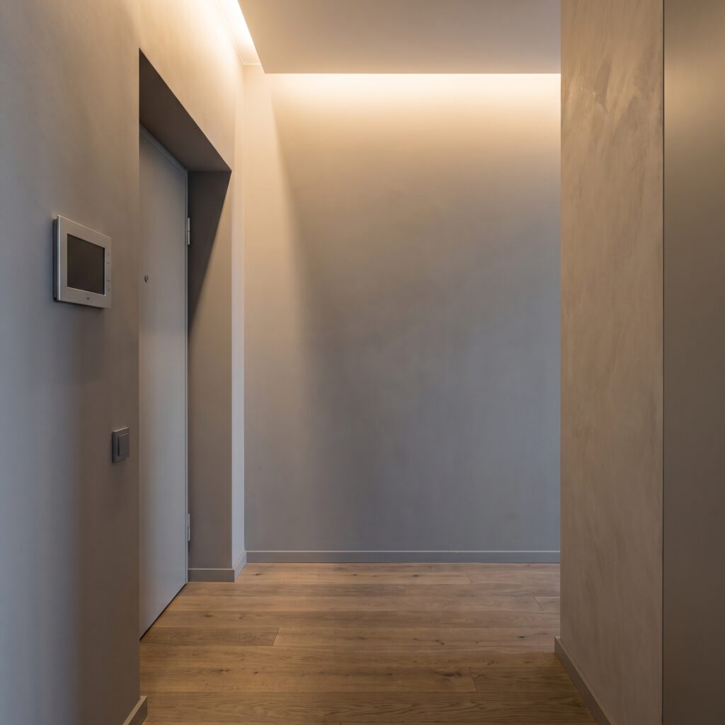 Korytarz przy drzwiach wejściowych do apartamentu z panelem wideodomofonu i czytnikiem zbliżeniowym na szarej ścianie, dębowa podłoga, punktowe ciepłe światło z sufitu.