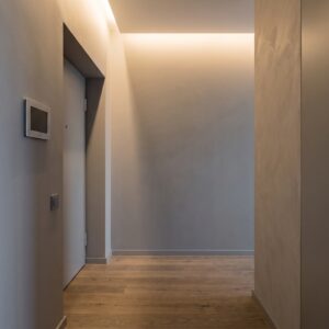 Korytarz przy drzwiach wejściowych do apartamentu z panelem wideodomofonu i czytnikiem zbliżeniowym na szarej ścianie, dębowa podłoga, punktowe ciepłe światło z sufitu.